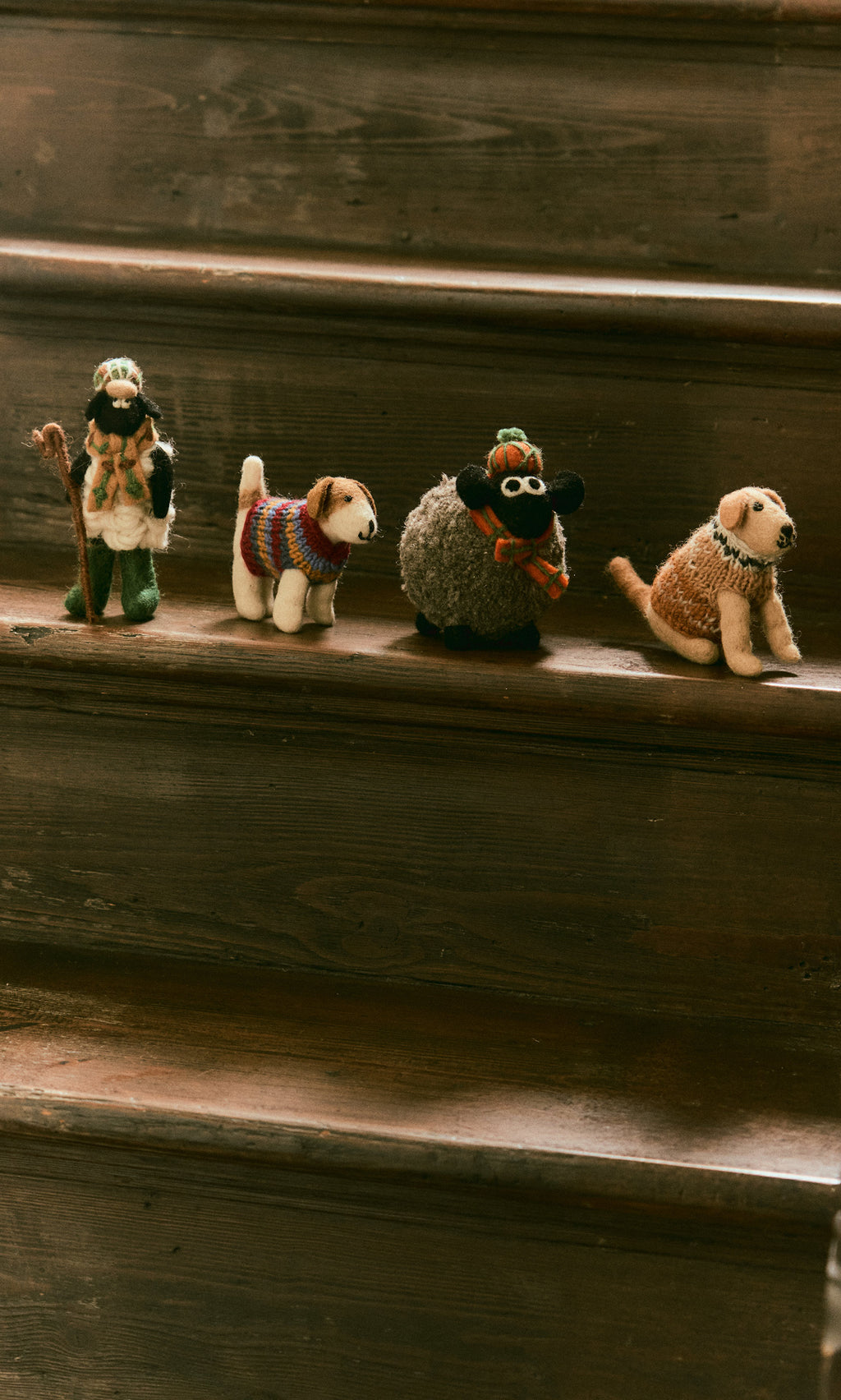 Plümo  Four handcrafted felted Flat Cap Sheep figurines wearing sweaters and hats lined up on a wooden staircase.