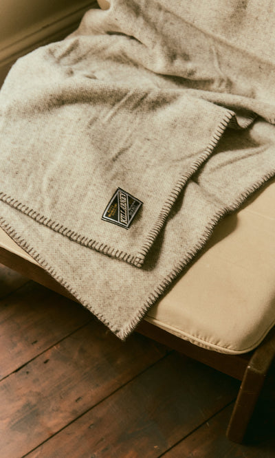 Plümo  The Kilkenny Blanket - Fossil is draped over a cushioned chair on a wooden floor, label visible.