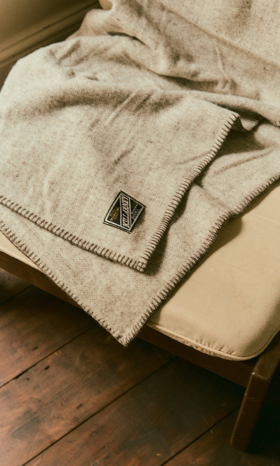 Plümo  The Kilkenny Blanket - Fossil is draped over a cushioned chair on a wooden floor, label visible.