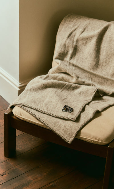 Plümo  Kilkenny Blanket - Fossil: pure new wool with stitched edge, draped on a cushioned wooden chair on wood floor.