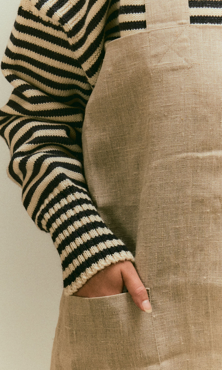Plümo  Person in a striped sweater and Linen Pinafore Apron with one hand tucked into the deep front pocket.
