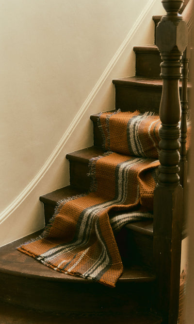 Plümo  The Random Throw drapes over dark wood stairs, its orange and gray stripes contrasting a white wall background.