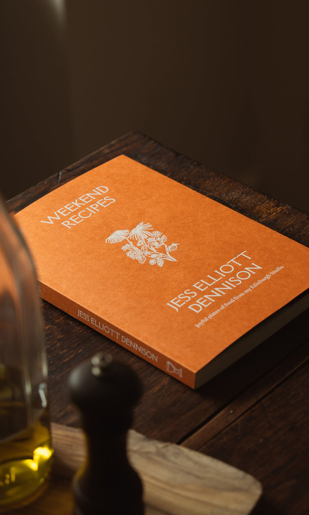 Plümo- The orange "Weekend Recipes by Jess Elliott Dennison" cookbook on a wooden table with oil and a pepper grinder.