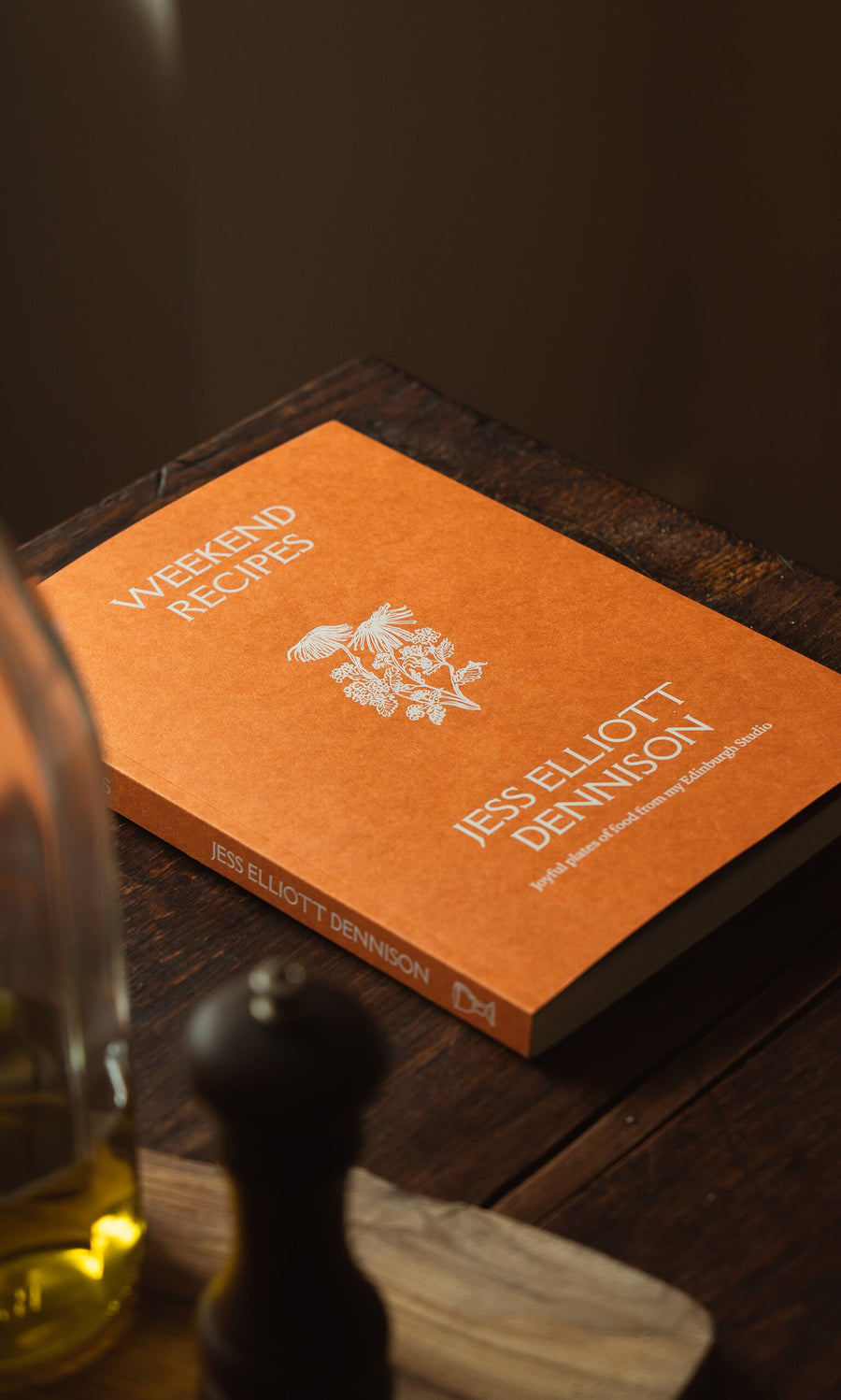 Plümo- The orange "Weekend Recipes by Jess Elliott Dennison" cookbook on a wooden table with oil and a pepper grinder.