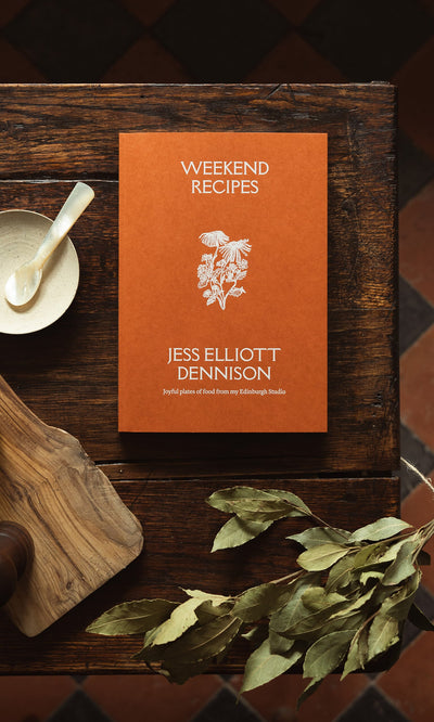Plümo- Weekend Recipes by Jess Elliott Dennison, an orange cookbook, ideal for weekends, sits on a wooden table with herbs.