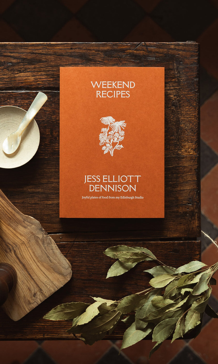 Plümo- Weekend Recipes by Jess Elliott Dennison, an orange cookbook, ideal for weekends, sits on a wooden table with herbs.