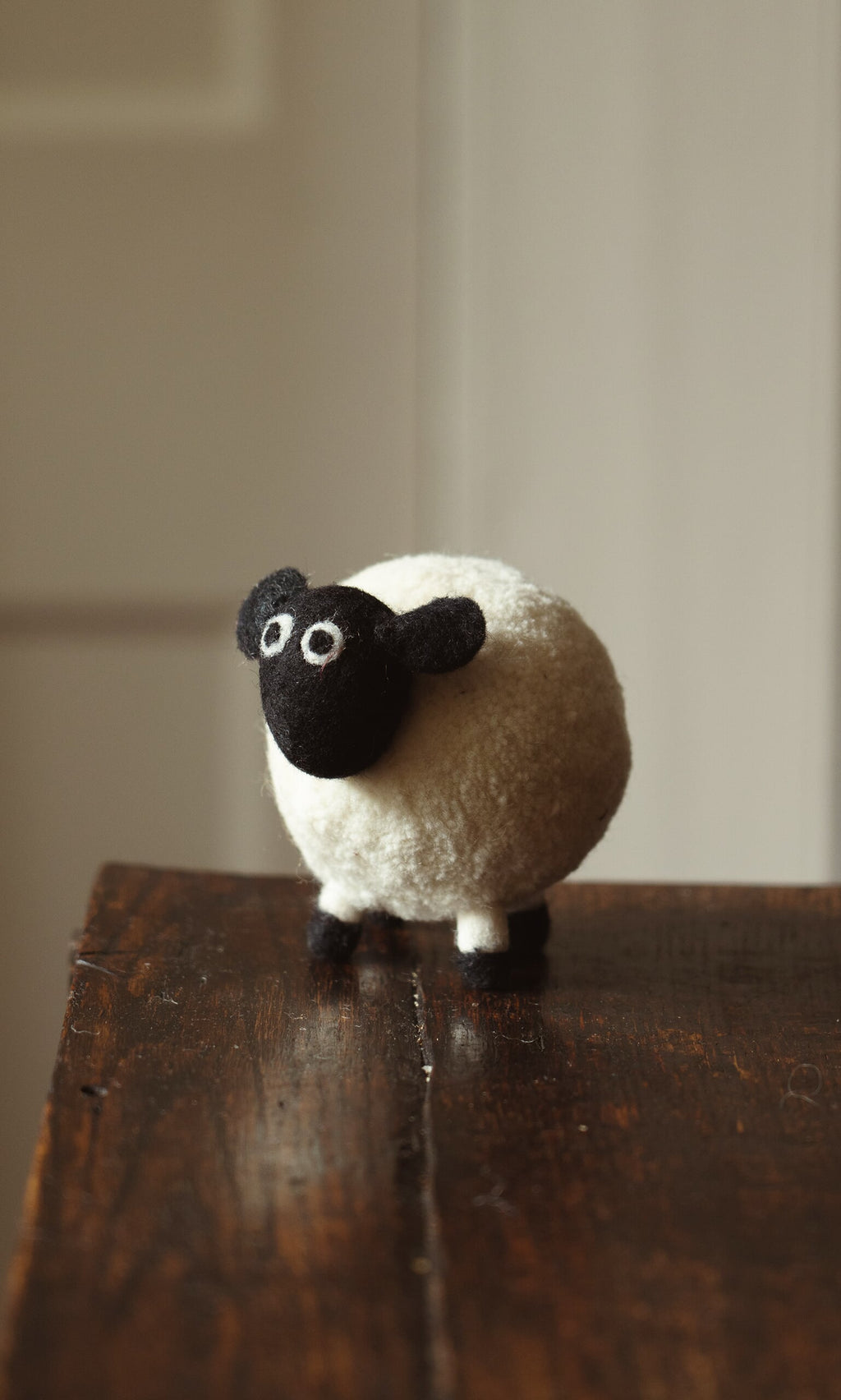 Plümo- Derek the Sheep, a small woolly figurine handmade in Nepal, stands on a wooden surface indoors.