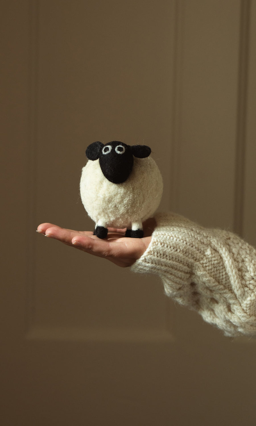 Plümo- A hand in a knit sweater holds Derek the Sheep, a small fairtrade hand-felted wool sheep, against a plain background.