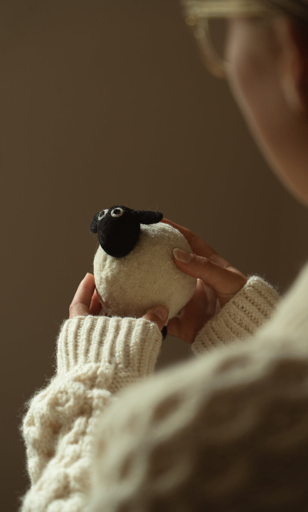 Plümo- Someone in a cream sweater holds Derek the Sheep, a small hand-felted wool toy with a black face and big eyes.