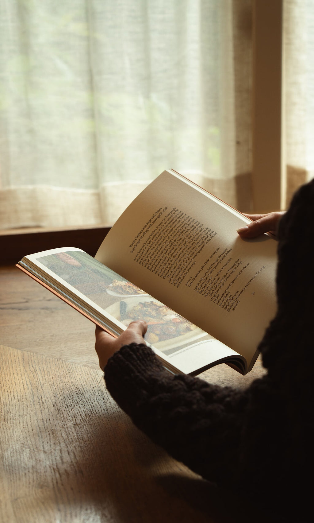 Plümo- Someone reads Weekend Recipes by Jess Elliott Dennison by a window with sheer curtains, sunlight streaming in.