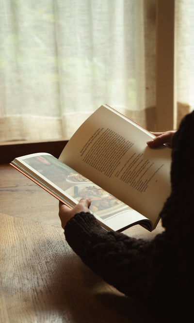 Plümo- Someone reads Weekend Recipes by Jess Elliott Dennison by a window with sheer curtains, sunlight streaming in.
