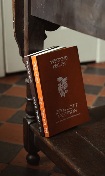 Plümo- The cookbook "Midweek Recipes By Jess Elliott Dennison" rests against a wooden bench on a tiled floor.