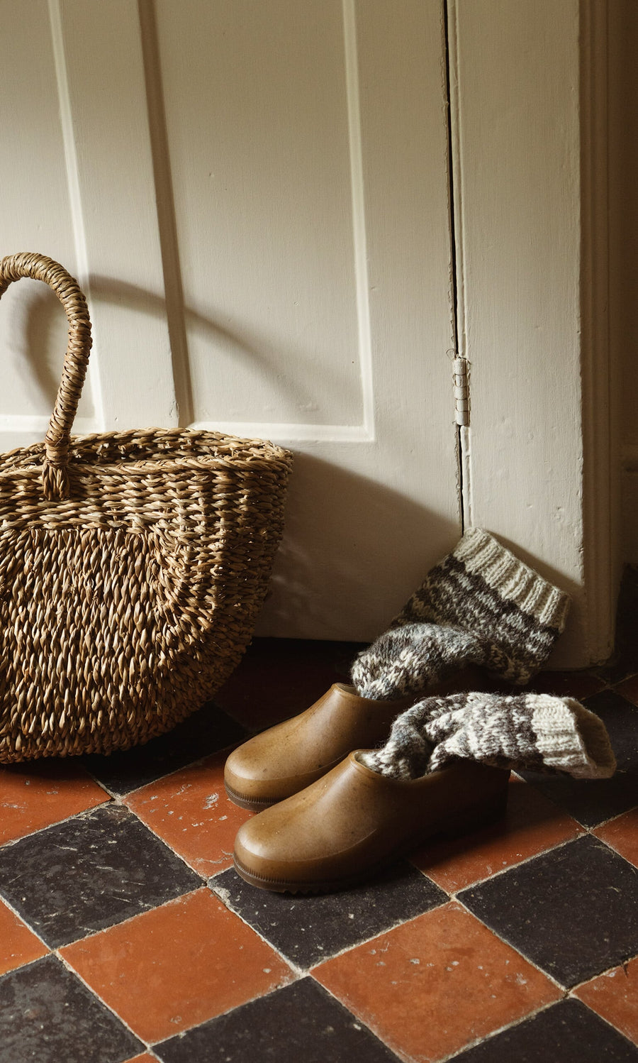Plümo- Hemp Clogs, a woven basket, and gray knit socks on a red and black tiled floor by a white door.