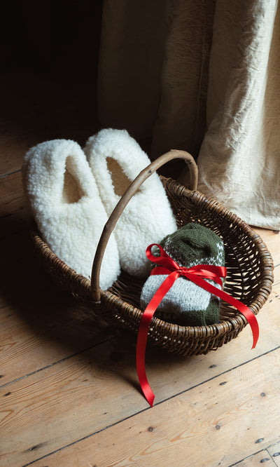Plümo- A wicker basket with Wool Slippers - Natural and knit socks tied with a red ribbon on a wooden floor.