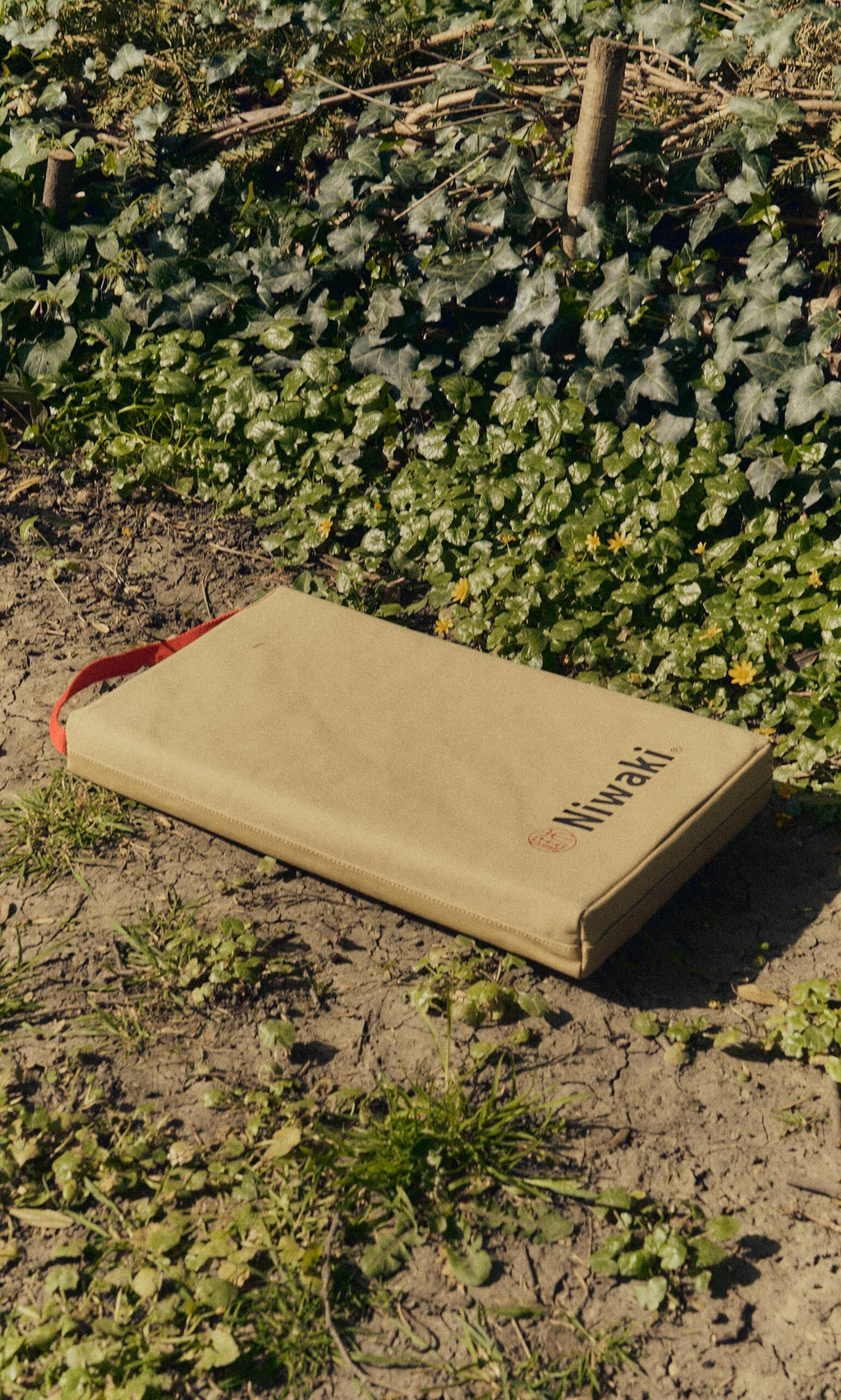 Plümo- The Niwaki Kneeler, tan with a red handle and logo, made of heavy-duty canvas, sits by leafy plants and yellow flowers.