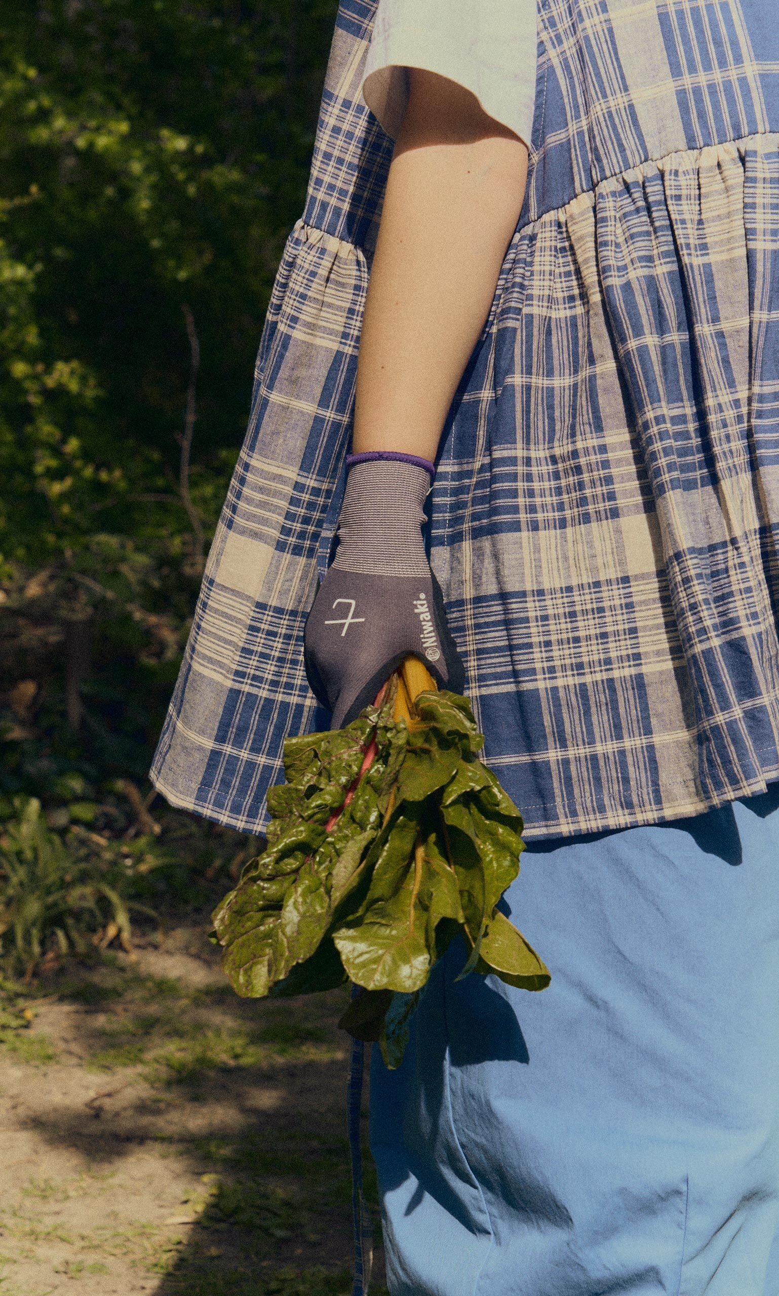 Plümo- Person in a plaid dress wears Niwaki gardening gloves, holding fresh leafy greens outdoors on a sunny day.