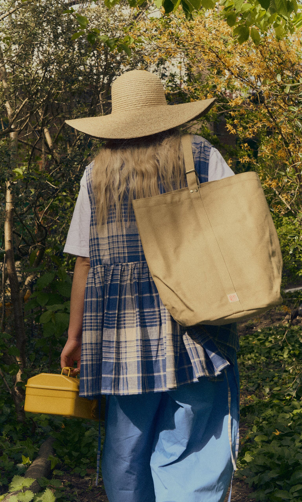 Plümo- A person in a straw hat and plaid vest carries a Canvas tool bag and yellow box through a sunlit, leafy forest.