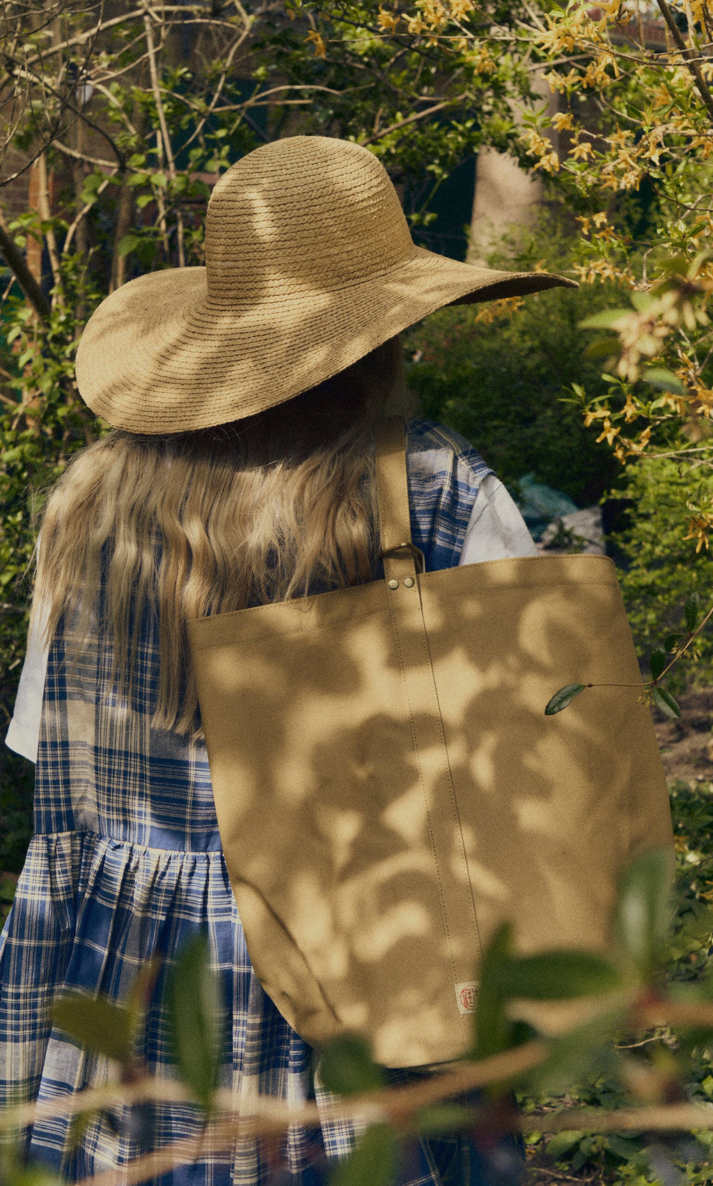 Plümo- Wearing a straw hat and plaid dress, she carries a Canvas tool bag among lush greenery outdoors.