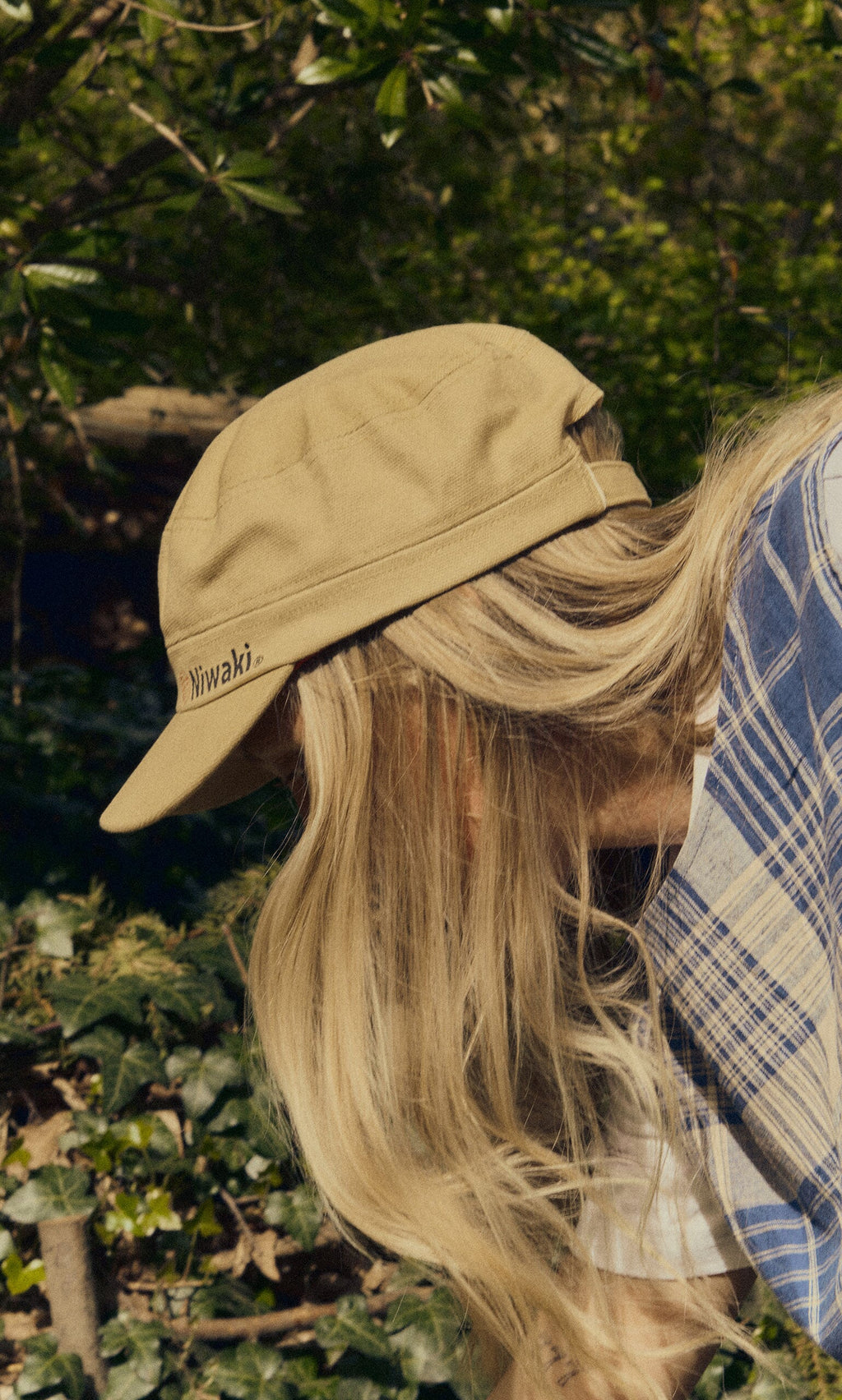 Plümo- Person with long blonde hair in a blue plaid shirt wearing a Niwaki Cap, bending over among green plants.