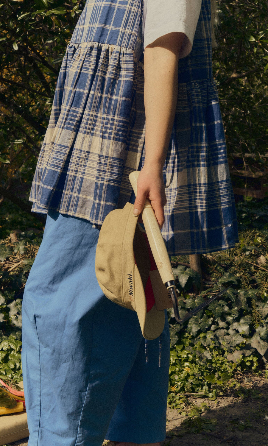 Plümo- Person in blue plaid dress and blue pants holds a Niwaki Cap and garden tool, standing outdoors near greenery.