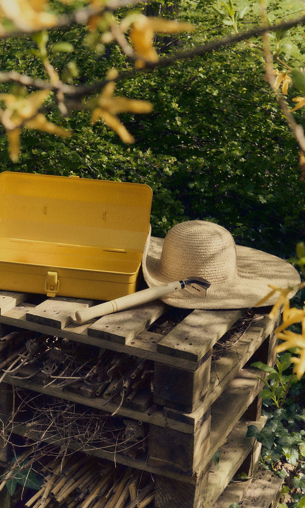 Plümo- A Small Weeding Hoe rests on stacked pallets among green foliage, near a straw hat and yellow toolbox.