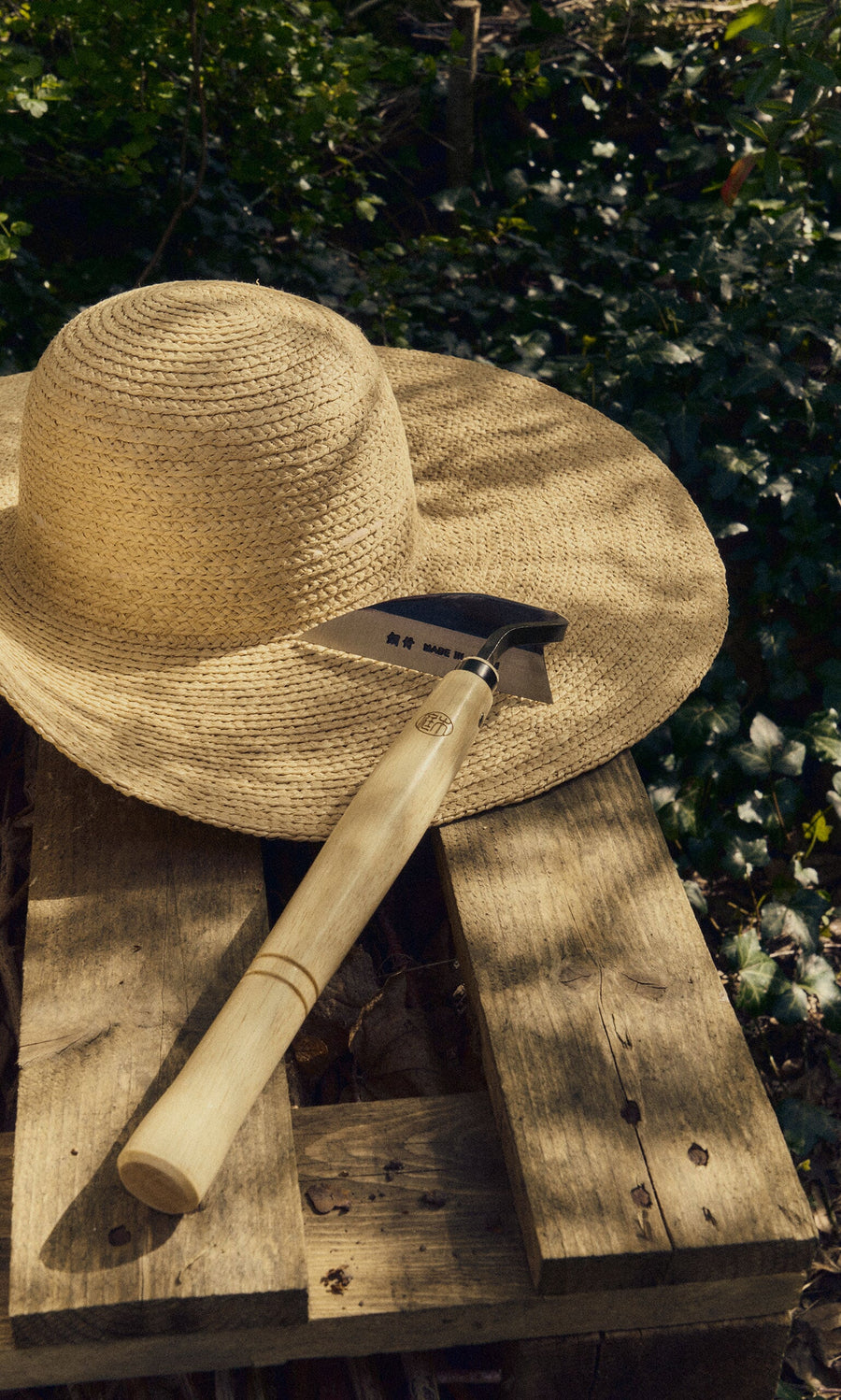 Plümo- A straw hat and a Small Weeding Hoe rest on a wooden pallet outdoors with greenery in the background.