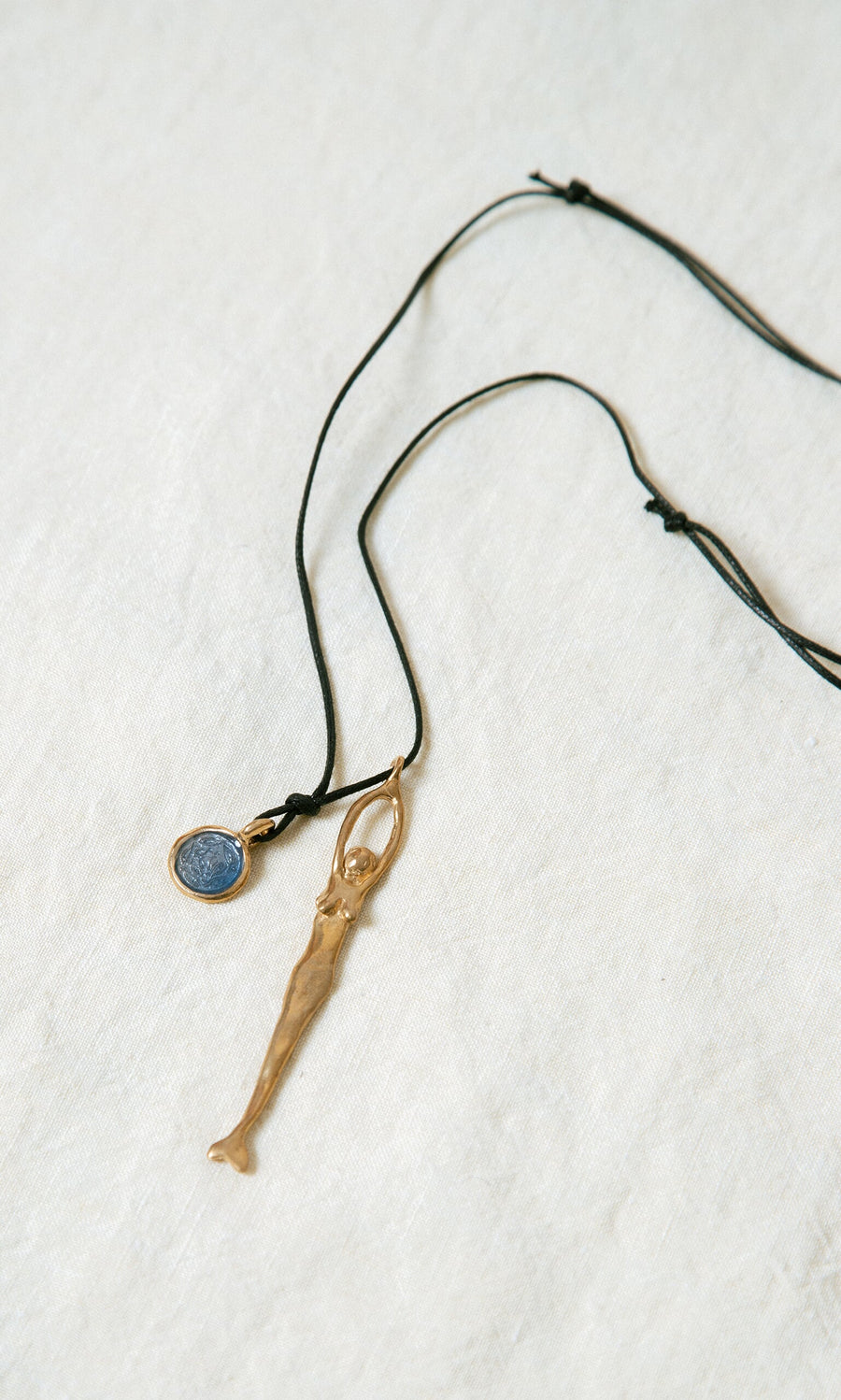 Plümo- The Capri Necklace features a black cord with a bronze mermaid pendant and blue ocean charm on white fabric background.