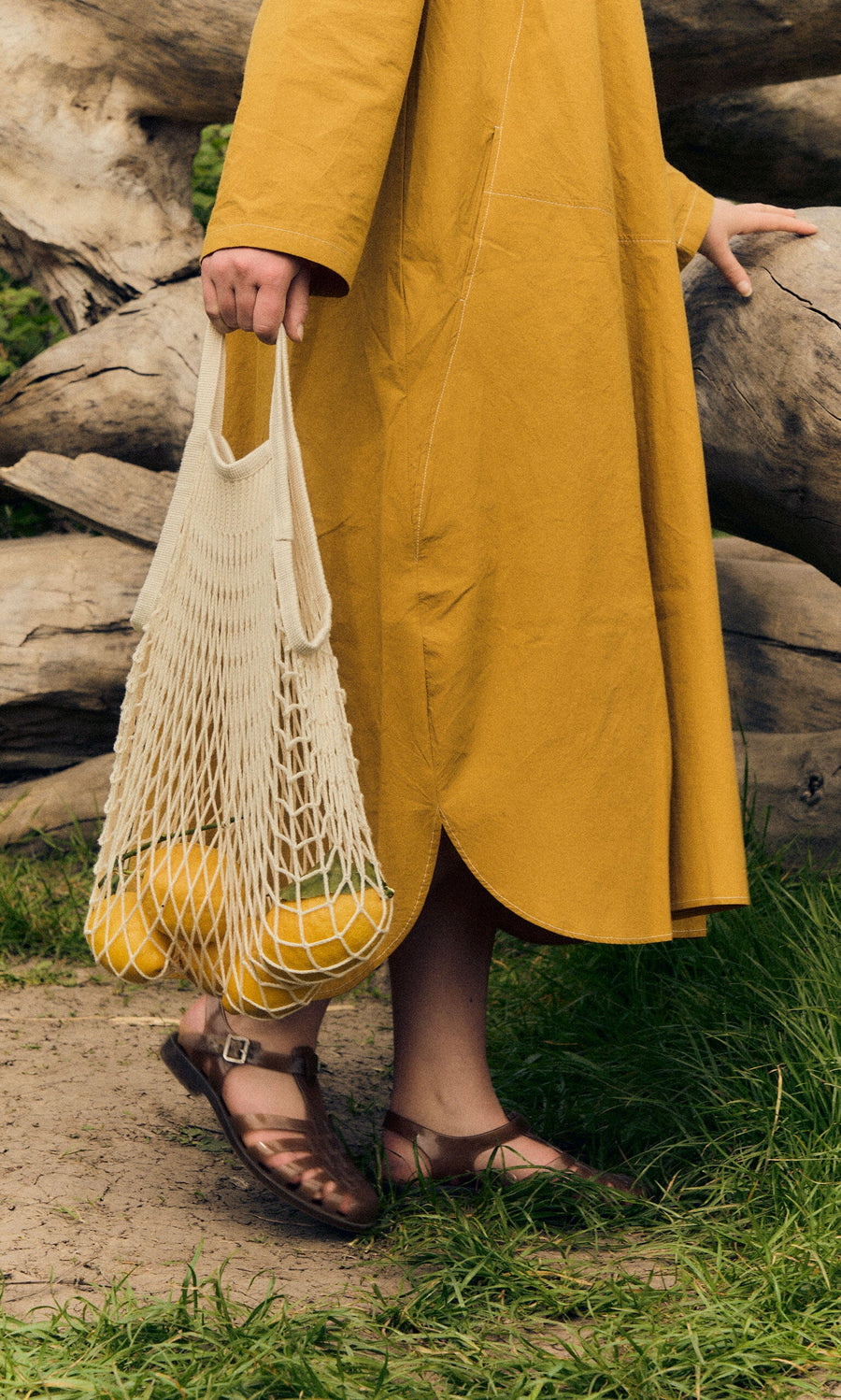 Plümo- Person in a yellow dress holds lemons in a mesh bag, wearing Hemp Sunchanvre Sandal, standing on grass by a log.