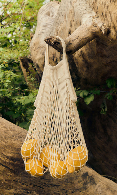 Plümo  A Net Shopping Bag in beige cotton, filled with lemons, hangs from an outdoor tree branch.