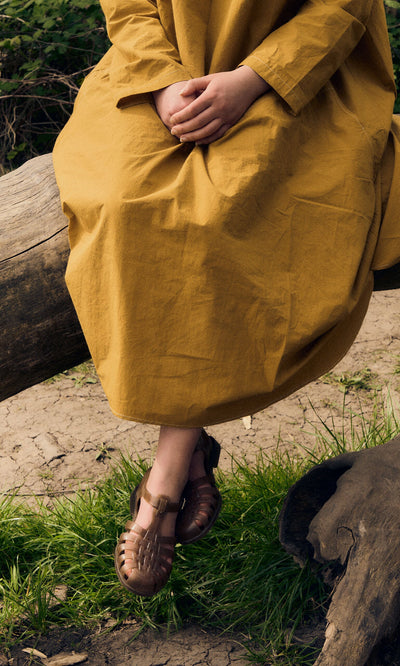Plümo- Person in a mustard yellow dress sits on a log, wearing brown Hemp Sunchanvre Sandals with hands folded outdoors.