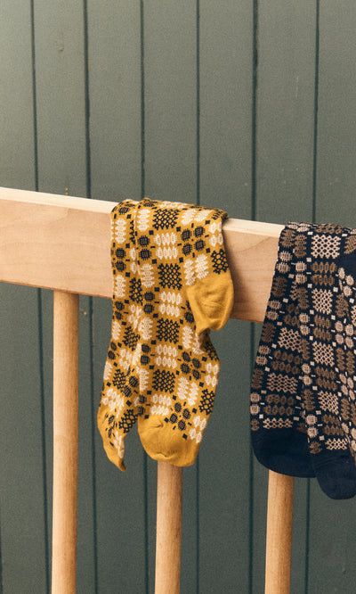 Plümo  Welsh Socks - Gold with tapestry pattern hang on a crib rail next to dark Italian cotton socks.