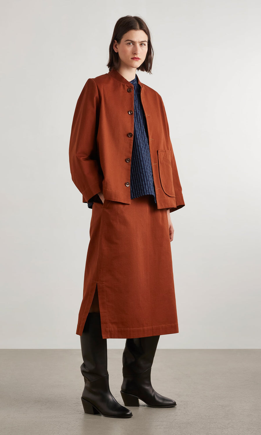 Plümo- A person in the rust Quinn Skirt and a navy top stands against a plain background in black heeled boots.