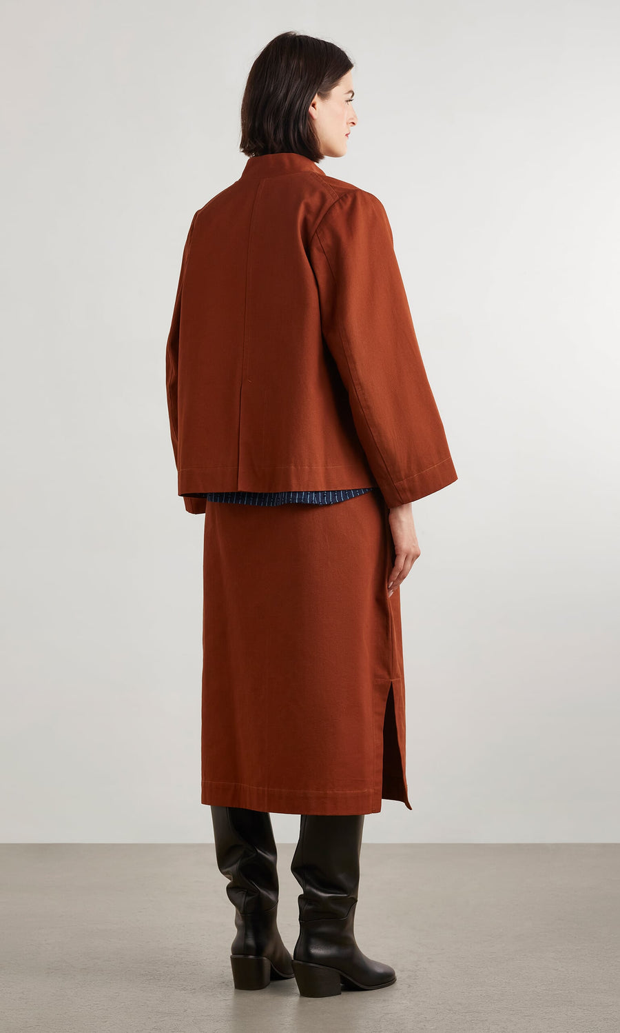 Plümo- A person in a rust-colored jacket and Quinn Skirt with a side slit, black boots, on a light gray floor.