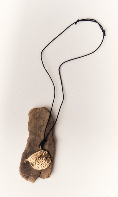 Plümo- A gold puffer fish pendant, crafted by lost-wax casting, hangs on a black cord necklace over driftwood.