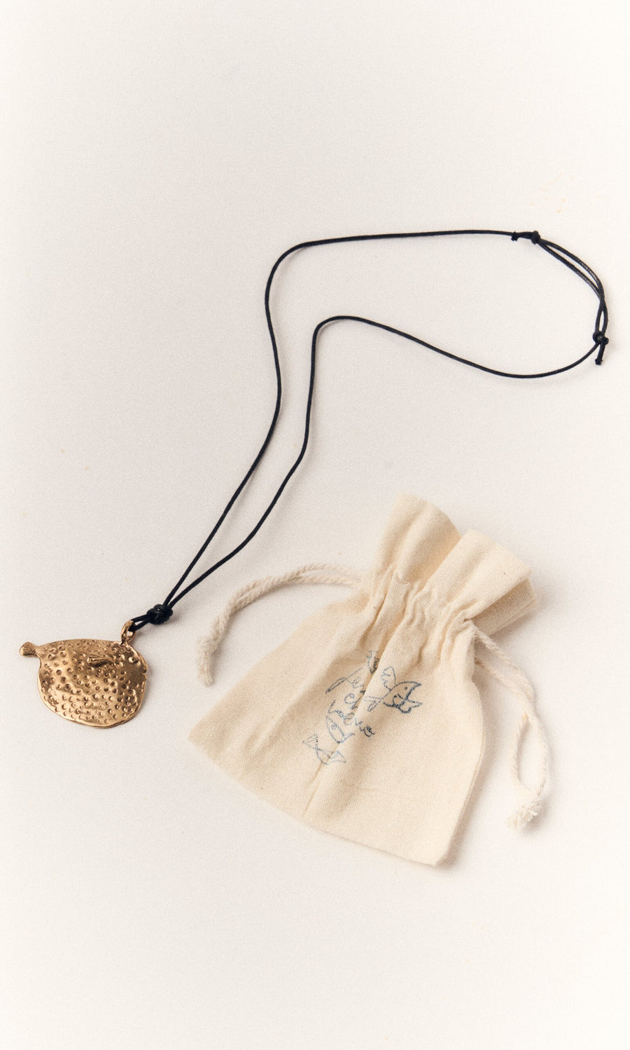 Plümo- The Puffer Fish Necklace features a handcrafted bronze puffer fish pendant on a black cord, shown with a cream pouch.