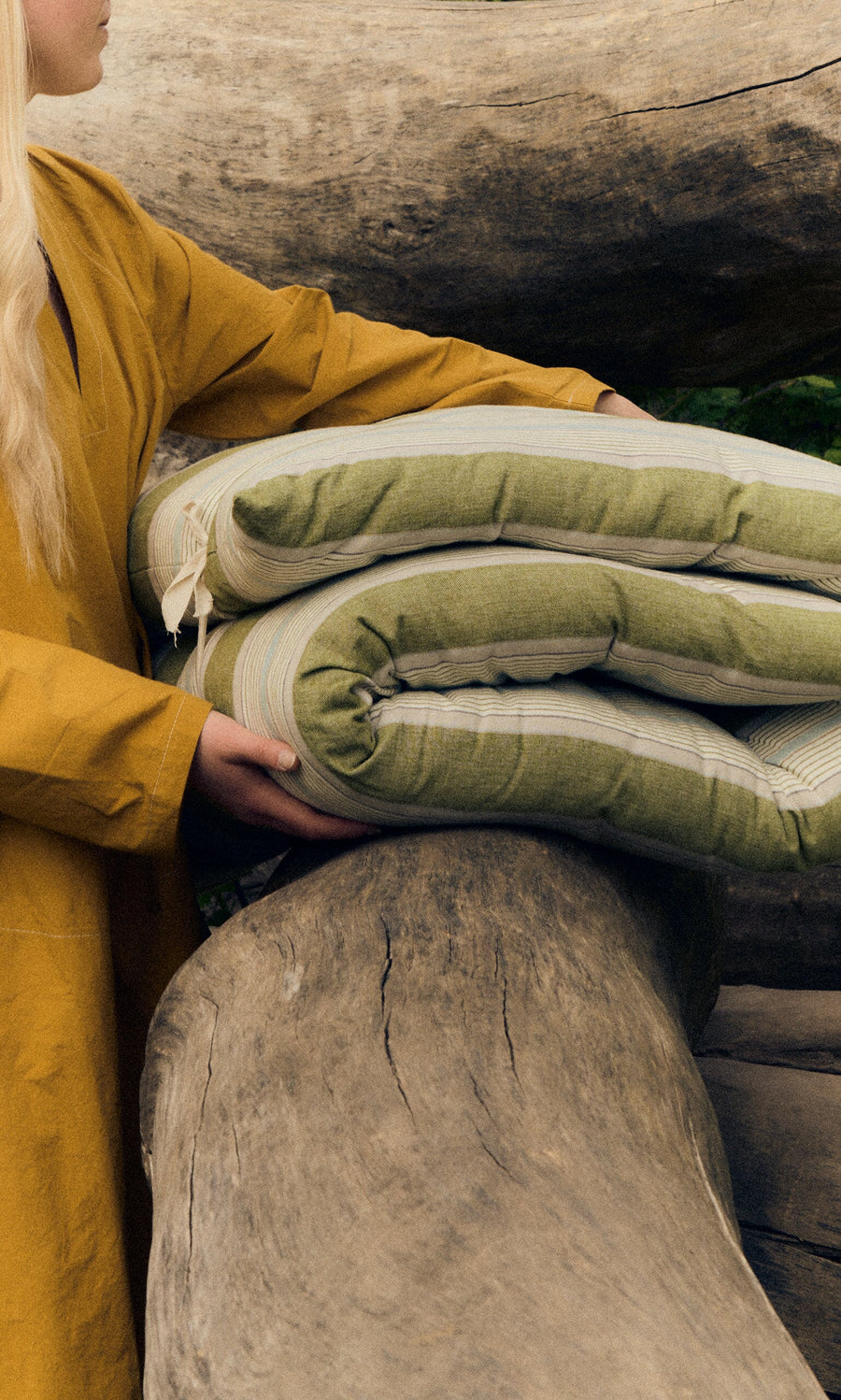 Plümo- A person in a mustard yellow coat holds a sustainable Quilted Mattress - Green Stripe on a large fallen tree trunk.
