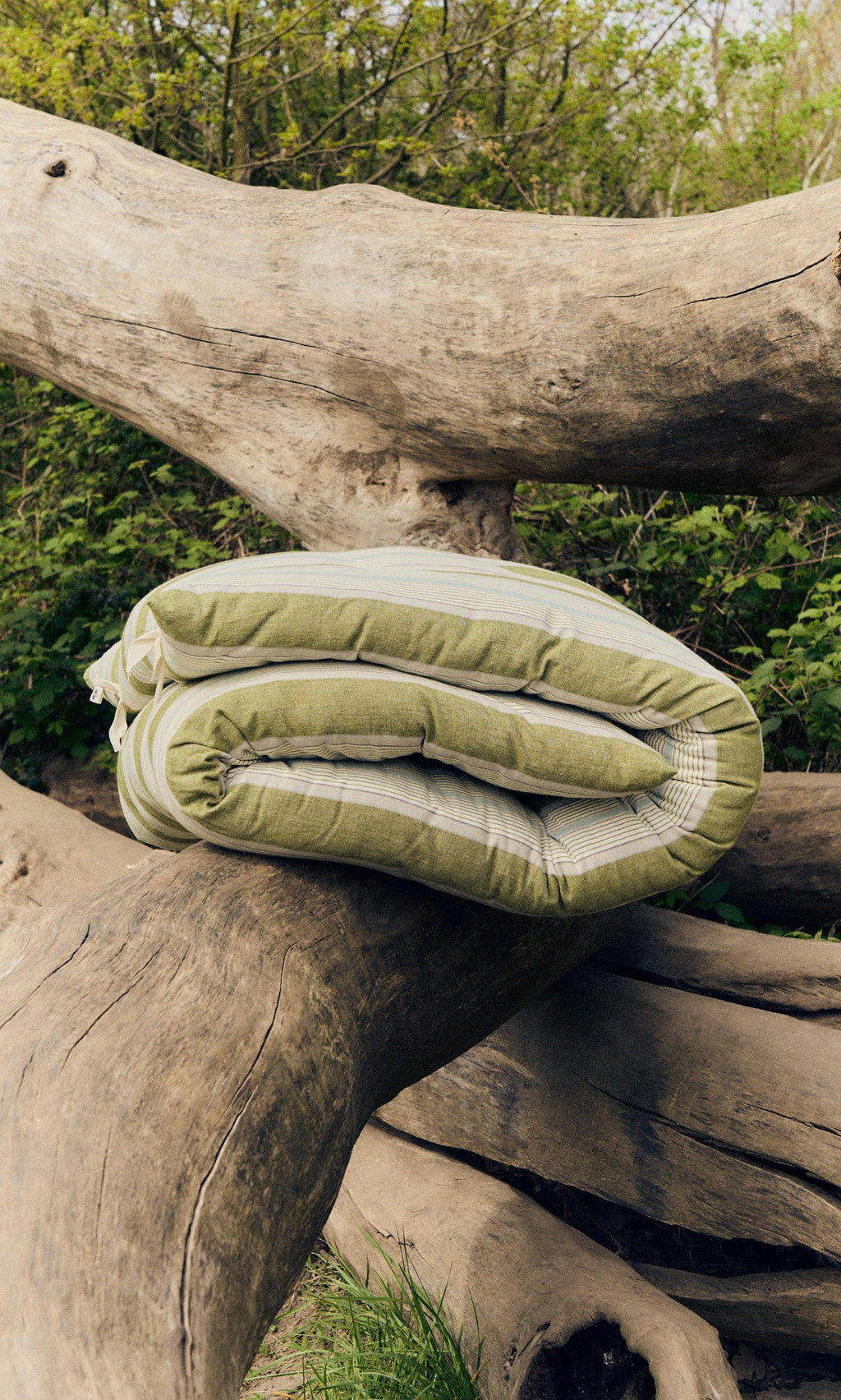 Plümo- The Quilted Mattress - Green Stripe, a sustainable foldable design, is displayed outdoors on tree trunks amid greenery.