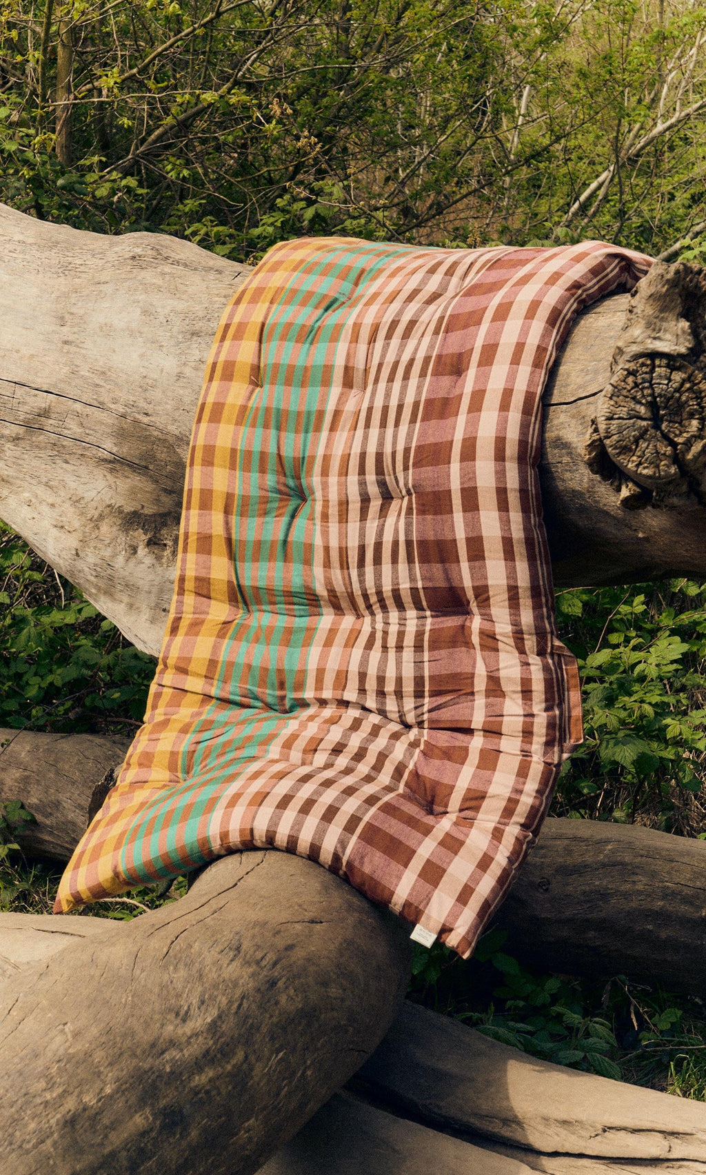Plümo- A Quilted Mattress - Red with a foldable design rests on large tree trunks in a wooded outdoor area.