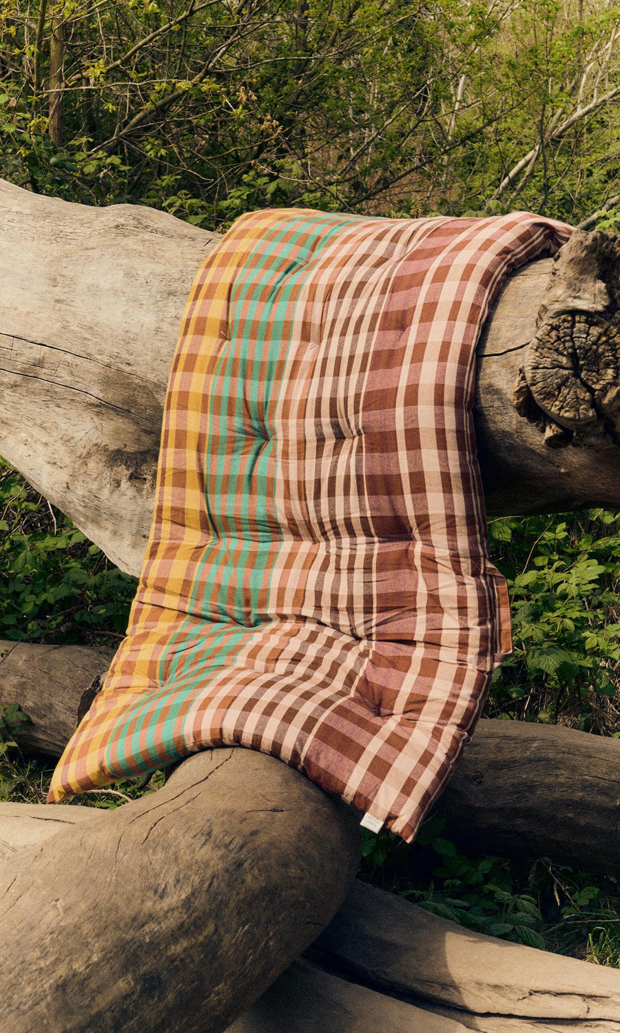 Plümo- A Quilted Mattress - Red with a foldable design rests on large tree trunks in a wooded outdoor area.