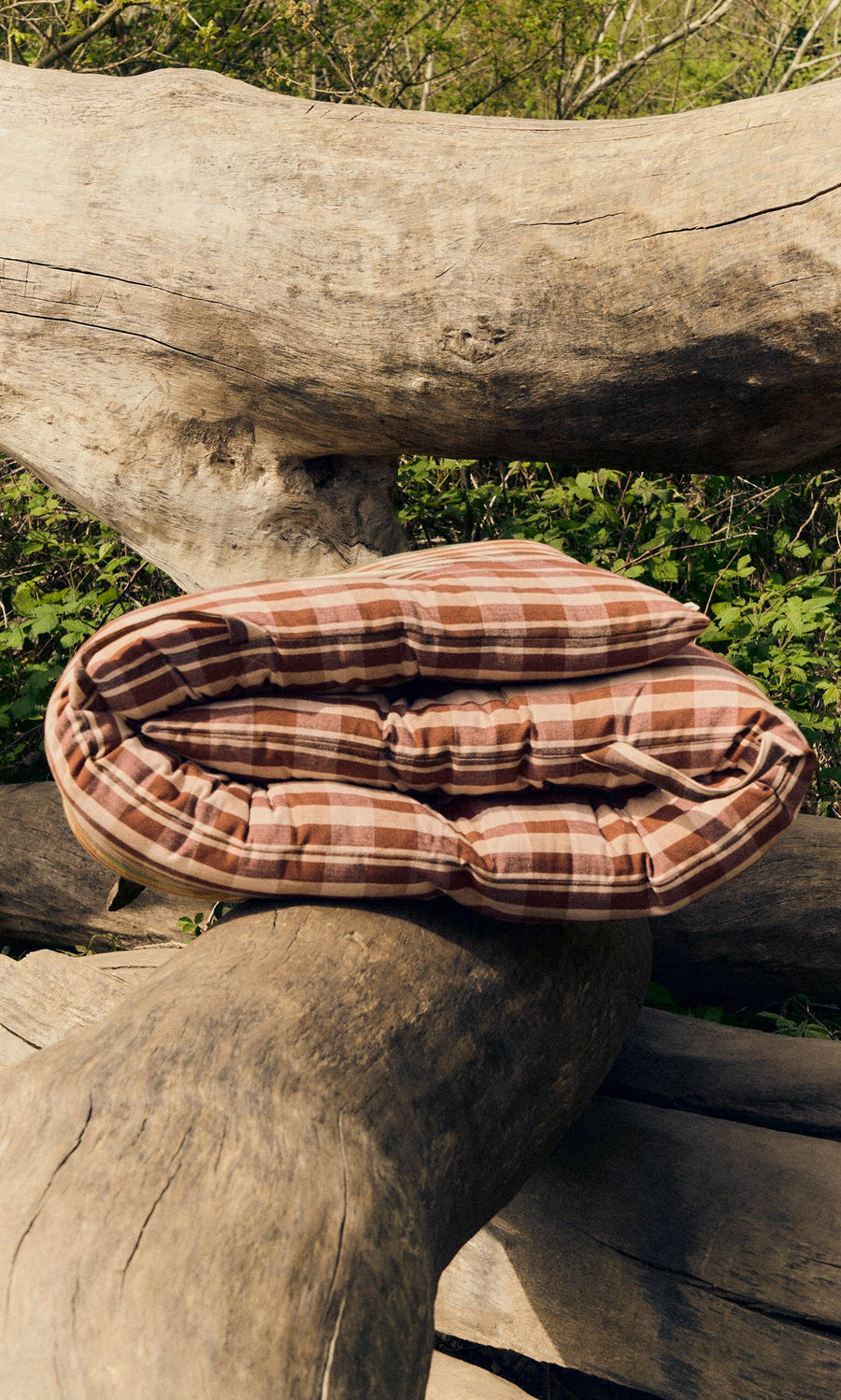 Plümo- A red quilted mattress with a sustainable, foldable design sits on large logs amidst greenery.