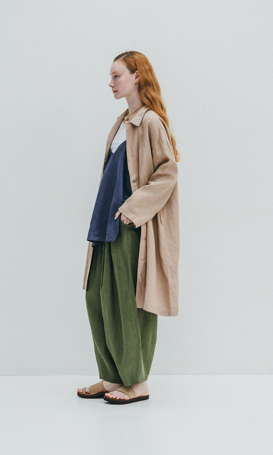 Plümo- A person in a beige coat, navy top, and green Quincy Pants stands sideways against a plain white background.