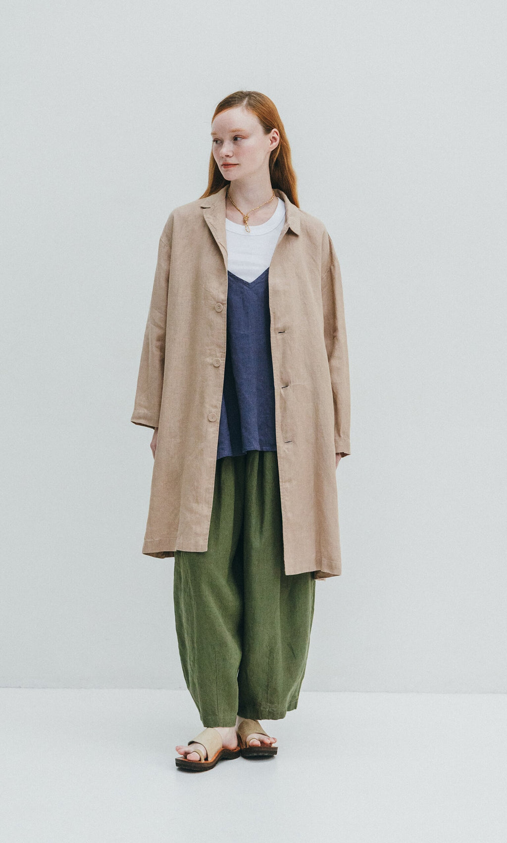 Plümo- Woman in green Quincy Pants with an elastic waist, a long beige coat, navy top, white shirt, and sandals.