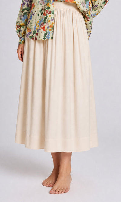 Plümo  Person wearing the Rika Wide Skirt, a flowy cream cotton skirt with elastic waist, standing barefoot in a floral blouse.
