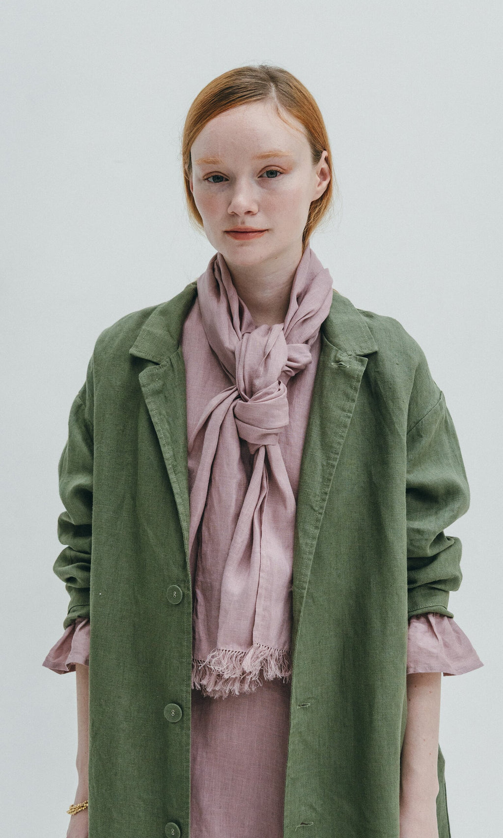Plümo- Young woman in a green coat wears the Rosie Scarf, a faded rose style with selvedge fringe, against a light background.