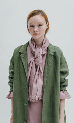 Plümo- Young woman in a green coat wears the Rosie Scarf, a faded rose style with selvedge fringe, against a light background.
