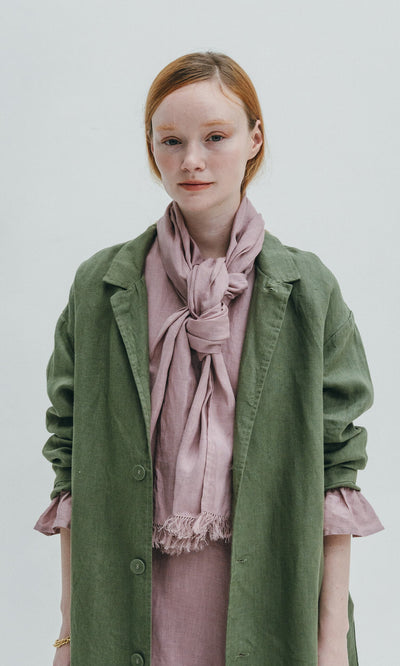 Plümo- Young woman in a green coat wears the Rosie Scarf, a faded rose style with selvedge fringe, against a light background.