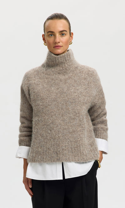 Plümo  Woman in Beel Rollneck sweater over white shirt, black pants, gold earrings, against a plain background.