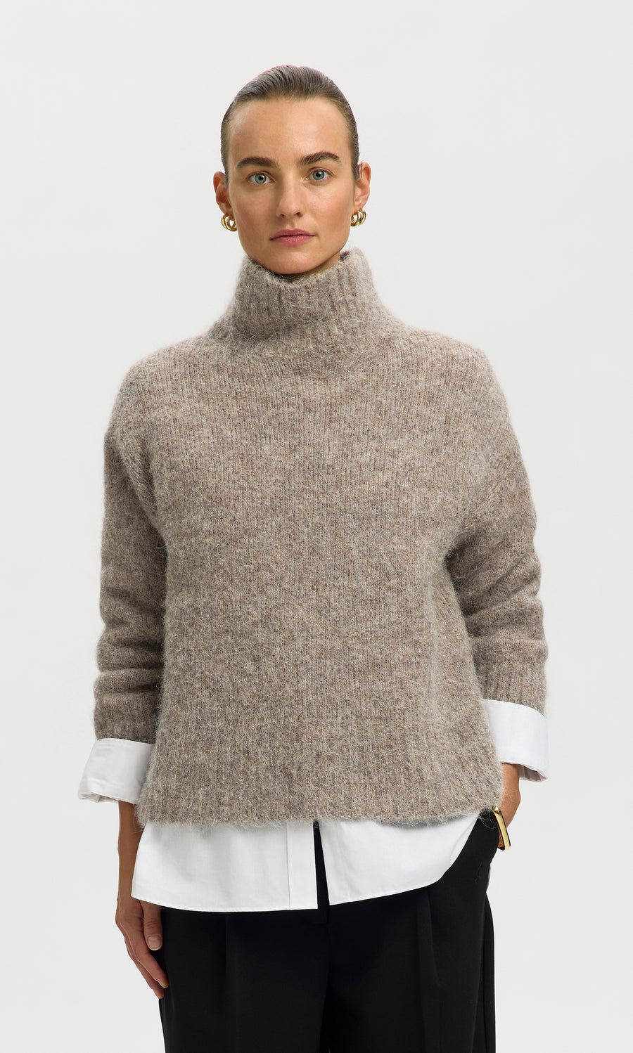 Plümo  Woman in Beel Rollneck sweater over white shirt, black pants, gold earrings, against a plain background.