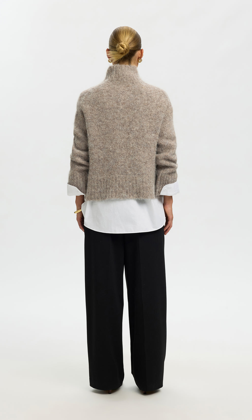 Plümo  Individual with a bun, in the Beel Rollneck beige sweater, white shirt, and black pants, stands facing away.
