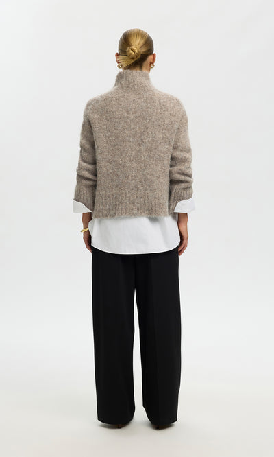Plümo  Individual with a bun, in the Beel Rollneck beige sweater, white shirt, and black pants, stands facing away.
